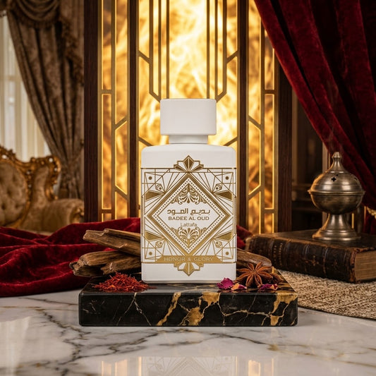 A white perfume bottle with ornate gold geometric detailing and Arabic text reading 'BADEE AL QUD' stands on a black marble base.