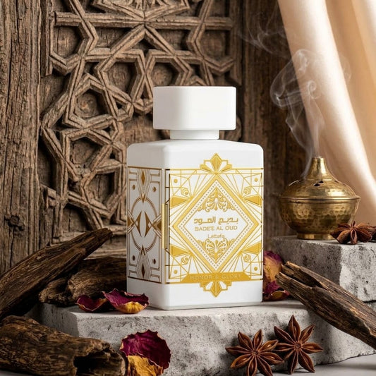 A white rectangular perfume bottle with gold geometric patterns and Arabic text, styled on stone blocks with dried wood, star anise, petals, a brass incense burner, and a carved wooden backdrop.