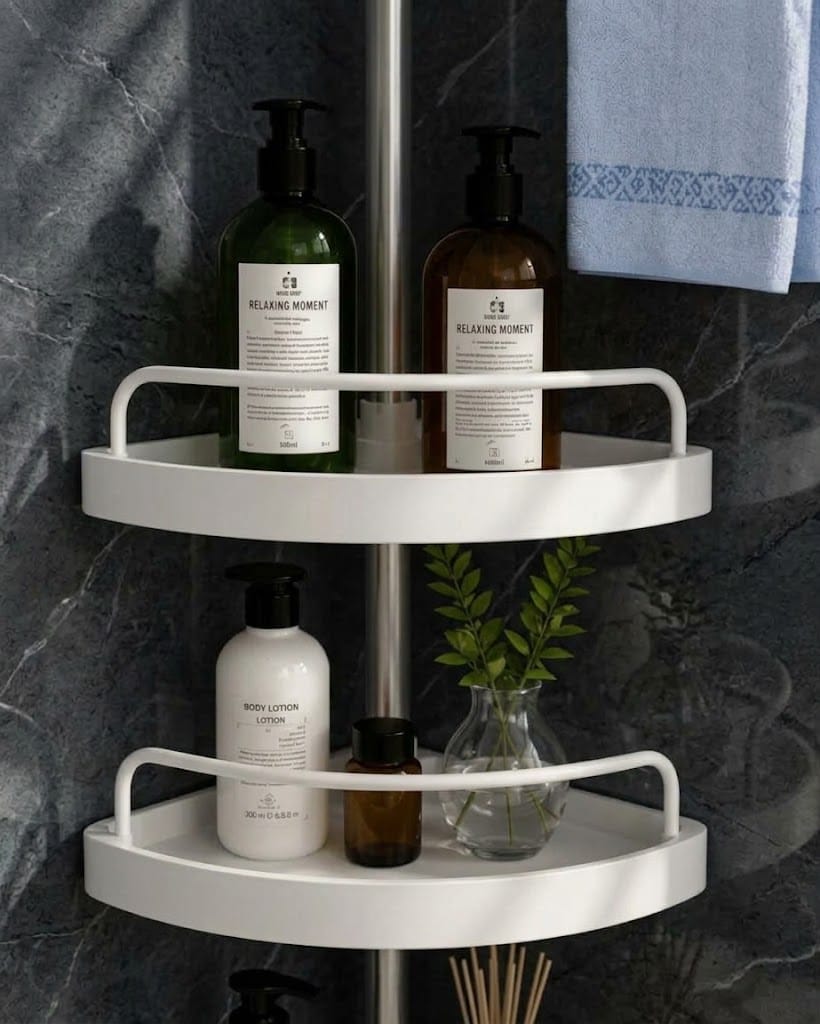 A white two-tier corner shower shelf mounted on a dark gray marble wall, styled with toiletries including green and white pump bottles, a small amber bottle, a glass vase with greenery, and a folded light blue towel.