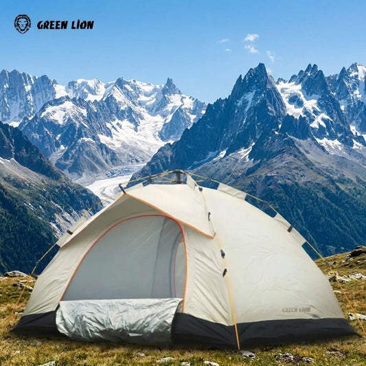 A dome-style camping tent pitched on a grassy mountain slope with sharp snow-capped peaks and blue sky in the background. The tent is light gray with dark gray base panels, orange trim, and mesh front door.