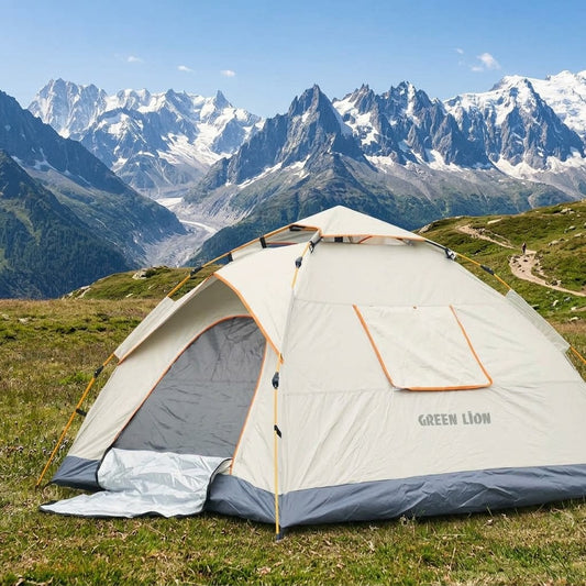 A Green Lion brand dome camping tent pitched on a grassy mountain meadow with snow-capped peaks in the background under a blue sky.