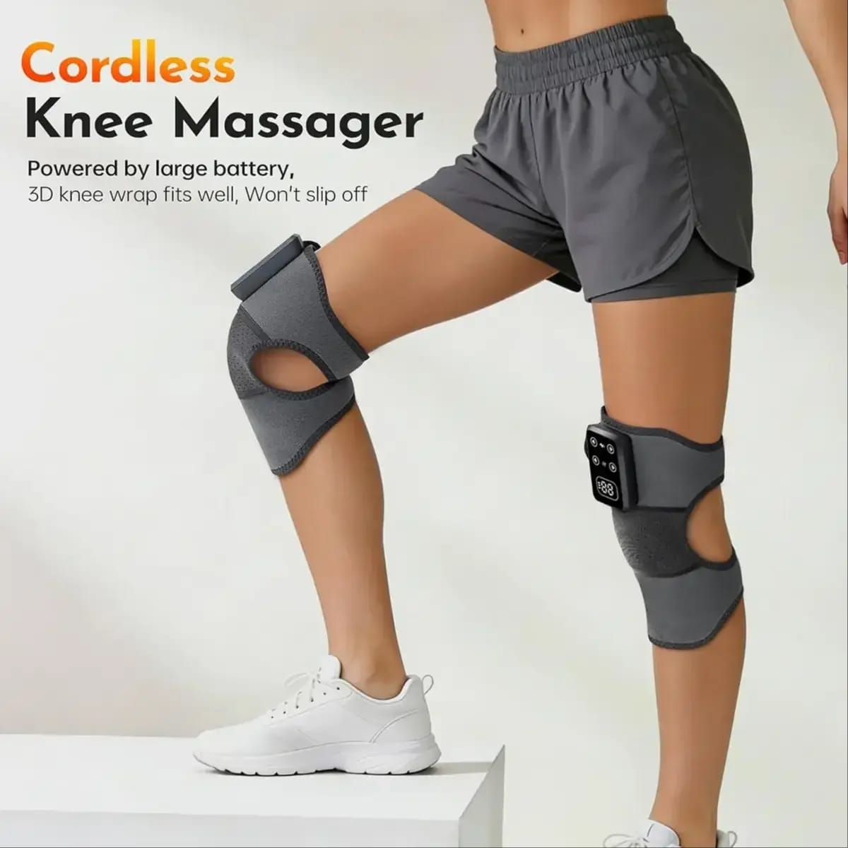 A person wearing gray 3D cordless knee massagers on both knees, paired with gray athletic shorts and white sneakers, standing on a white platform against a light background. 'Cordless Knee Massager' with feature copy below.