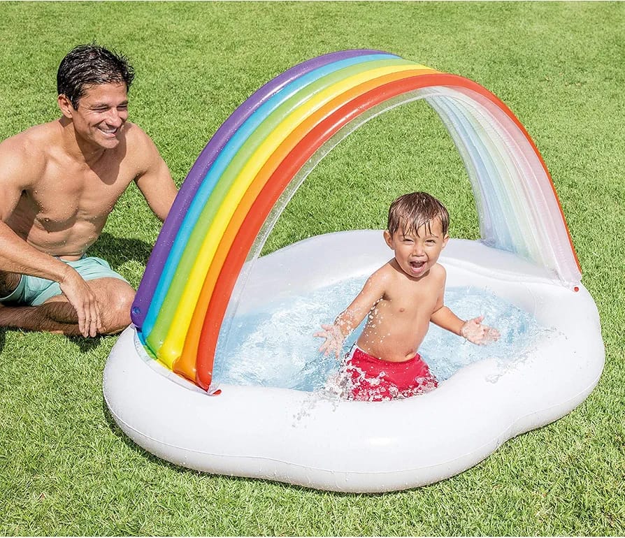 Child playing in a inflatable pool with rainbow arch and canopy, surrounded by grass.