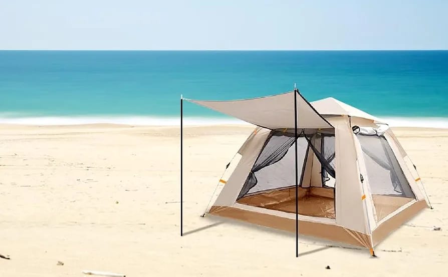 A beige and brown beach tent with an attached white canopy tarp is pitched on a sandy beach with the blue ocean and sky in the background.