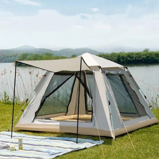 A large beige and gray outdoor camping tent with a pitched roof and mesh windows, set on grass beside a lake with hills in the background. A striped blanket and a bottle are visible in the foreground.