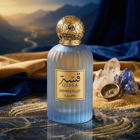 A ribbed frosted light blue perfume bottle with a gold label reading QISSA EMPOWER VALLEY CALIPH, topped with an ornate gold cap. The bottle is styled on a reflective dark marble surface with gold sand, blue velvet, a pocket watch, crystals, and a scenic mountain landscape backdrop.
